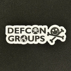 May include: White sticker with black text that reads "DEFCON GROUPS" with a skull and crossbones with a smiley face on the left side and a skull and crossbones with a smiley face on the right side.