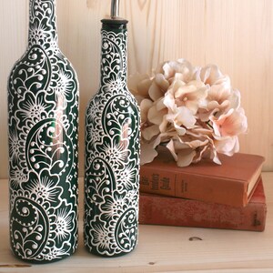 May include: Two green glass bottles with white floral designs and stainless steel spouts. The bottles are decorated with intricate white patterns. A bouquet of pink flowers and two books are in the background.
