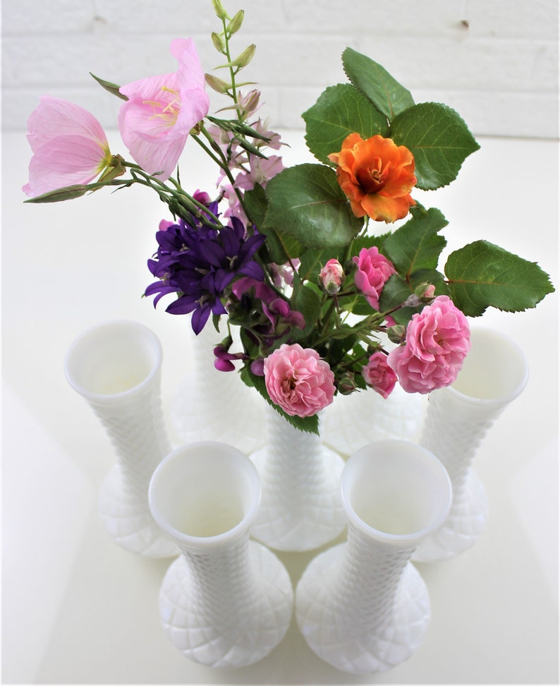 Puede incluir: Seis jarrones de vidrio de leche blanco con un patr&oacute;n de diamante texturizado. Los jarrones est&aacute;n dispuestos en c&iacute;rculo con un ramo de flores rosas, naranjas y moradas en el centro.