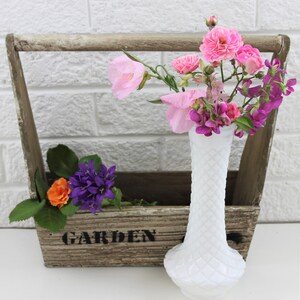 Puede incluir: Un jarr&oacute;n de vidrio de leche blanco con un patr&oacute;n de diamante contiene un ramo de flores rosas y moradas. El jarr&oacute;n est&aacute; sentado en una caja de madera con la palabra "JARDIN" pintada en la parte delantera.