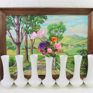 Puede incluir: Ocho jarrones de vidrio de leche blanco con un patr&oacute;n de diamante texturizado. Los jarrones est&aacute;n dispuestos en una fila frente a una pintura enmarcada de un paisaje campestre.