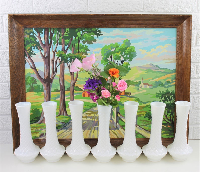 Puede incluir: Ocho jarrones de vidrio de leche blanco con un patr&oacute;n de diamante texturizado. Los jarrones est&aacute;n dispuestos en una fila frente a una pintura enmarcada de un paisaje campestre.