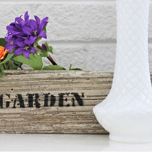 Puede incluir: Un jarr&oacute;n de vidrio de leche blanco con un dise&ntilde;o texturizado se encuentra junto a una maceta de madera con la palabra "JARD&Iacute;N" pintada en ella. Hay flores moradas y naranjas en la maceta.