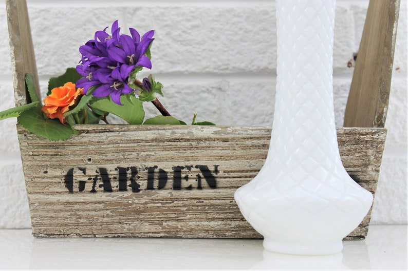 Puede incluir: Un jarr&oacute;n de vidrio de leche blanco con un dise&ntilde;o texturizado se encuentra junto a una maceta de madera con la palabra "JARD&Iacute;N" pintada en ella. Hay flores moradas y naranjas en la maceta.