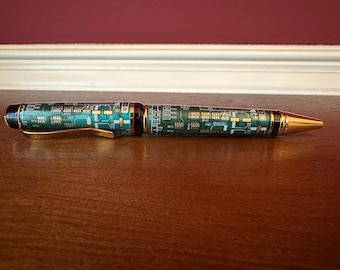 Könnte beinhalten: Ein Kugelschreiber mit Leiterplatten-Design. Der Stift hat ein grünes und goldenes Muster mit goldenen Akzenten an Clip und Spitze. Der Stift liegt auf einer Holzoberfläche.