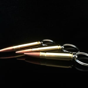 May include: Three gold-colored bullet casings with silver key rings attached. The casings are arranged in a row, with the top casing slightly offset to the left. The casings are on a black surface.