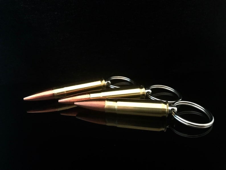 May include: Three gold-colored bullet casings with silver key rings attached. The casings are arranged in a row, with the top casing slightly offset to the left. The casings are on a black surface.