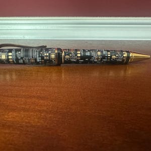 Könnte beinhalten: Ein schwarz-goldener Stift mit Leiterplattenmuster. Der Stift hat eine goldfarbene Spitze und Akzente sowie einen Clip. Der Stift liegt auf einer Holzoberfläche.