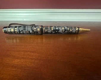 Könnte beinhalten: Ein schwarz-goldener Stift mit Leiterplattenmuster. Der Stift hat eine goldfarbene Spitze und Akzente sowie einen Clip. Der Stift liegt auf einer Holzoberfläche.