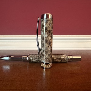 May include: A luxury pen with a snakeskin pattern on the barrel and cap. The pen has silver-colored metal accents and a gold-colored band near the base of the cap. The pen is lying on a wooden surface.