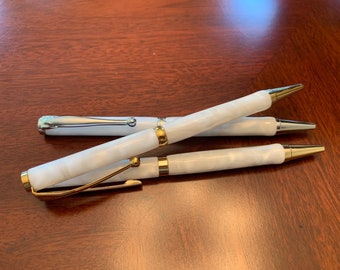 May include: Three ballpoint pens with white marbled barrels and metallic accents. Two pens feature gold-tone clips, bands, and tips, while one pen features silver-tone clips, bands, and tips. The pens are arranged in a stacked, overlapping formation.