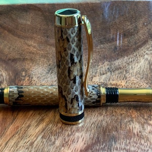 Könnte beinhalten: Ein goldener und schwarzer Stift mit Schlangenhautmuster. Der Stift liegt auf einer Holzoberfläche.