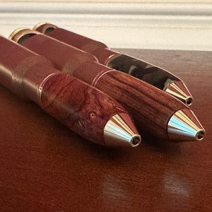 May include: Three wooden pens with metal tips, each with a different wood grain pattern. The pens are arranged in a row, with the tips pointing to the right.