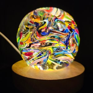 May include: A colorful glass orb with swirling patterns of red, yellow, blue, green, and white. The orb is illuminated from within and sits on a wooden base.