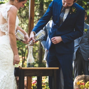 May include: A wedding ceremony with a bride and groom pouring colored liquids from glass bottles into a larger glass vase on a wooden table. The bride is wearing a white lace dress and the groom is wearing a blue suit.