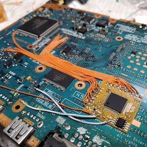 May include: A close-up of a circuit board with a small, square circuit board attached. Orange wires connect the two boards. The main board is teal with various chips and components. Text on the board includes "Sony Computer Entertainment".