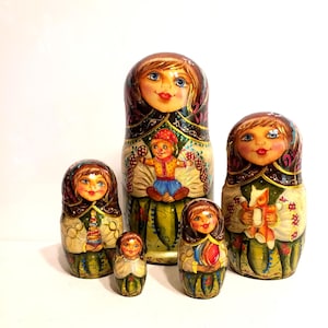 May include: A set of five hand-painted wooden nesting dolls featuring figures with brown hair and green-toned clothing. The largest doll displays a child figure on its front, while the others hold small toys like a stacker, a ball, and a fox.