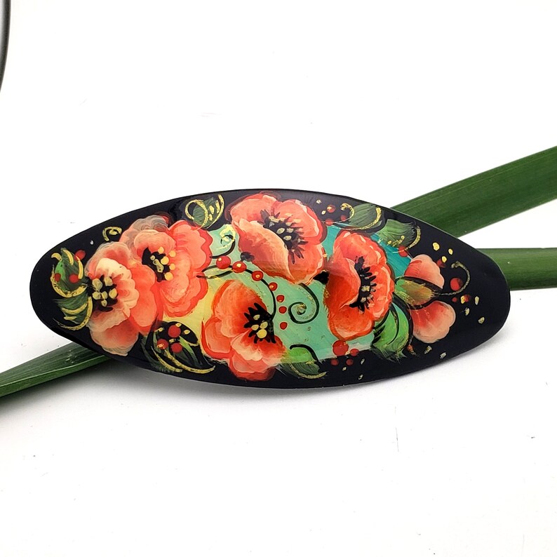 May include: Black oval-shaped hair clip with a hand-painted design of red and orange flowers on a teal background.