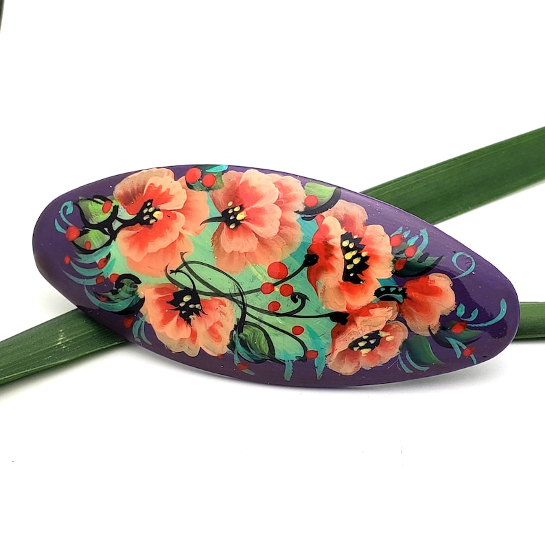 May include: Purple oval-shaped hair clip with a hand-painted floral design. The design features red, orange, and yellow flowers with green leaves and stems on a purple background.
