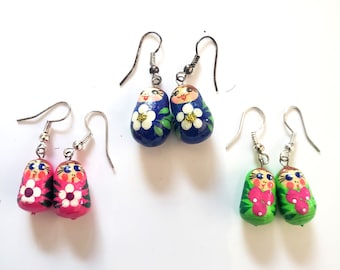 May include: Three pairs of Matryoshka doll earrings. One pair is pink with white flower accents, another is blue with white flower accents, and the last is green with pink flower accents. Each doll has a smiling face and silver-tone ear wires.
