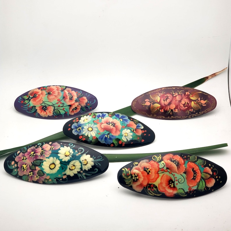 May include: Five oval-shaped hair clips with hand-painted floral designs. The clips are black with a variety of colorful flowers, including red, pink, yellow, and blue.