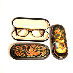 Puede incluir: Gafas con montura negra dentro de un estuche abierto negro y crema. También hay dos estuches decorativos, negros, de forma ovalada, para gafas con pinturas ornamentadas y coloridas. Un estuche presenta un diseño de fénix, el otro una figura.