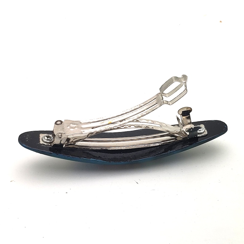 May include: A silver hair clip with a blue and black oval-shaped base. The clip is open and ready to be used.