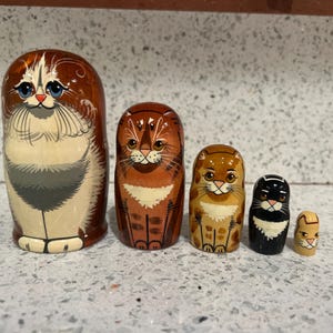 May include: A set of five wooden nesting dolls, each painted with a different cat design. The largest doll is brown and white, while the others are various shades of brown, black, and tan. The dolls are arranged in a row.