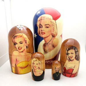 May include: A set of five wooden nesting dolls featuring images of Marilyn Monroe. The largest doll has a red, white, and blue design with a portrait of Monroe. The other dolls have different portraits of Monroe in various outfits.