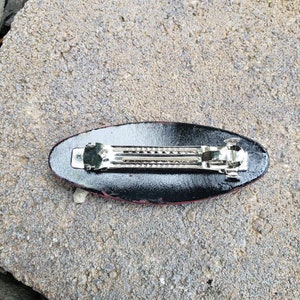 May include: Black and red oval hair clip with a silver metal clasp. The clip is designed to hold hair in place.