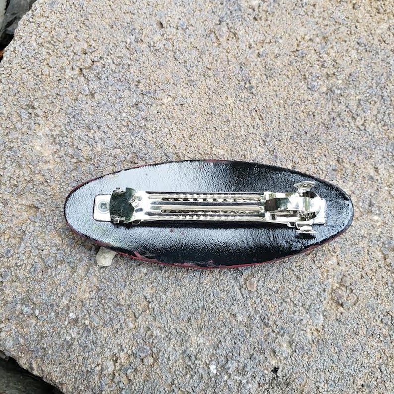May include: Black and red oval hair clip with a silver metal clasp. The clip is designed to hold hair in place.