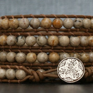 May include: A multi-layered leather wrap bracelet with brown leather cord and beige and brown speckled beads. A silver charm with a Celtic knot design is attached to the bracelet.
