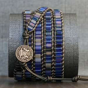 Puede incluir: Un brazalete de cuero de m&uacute;ltiples hebras con cuentas de vidrio azul y gris y un dije de moneda de plata. El brazalete est&aacute; envuelto alrededor de un cilindro negro.