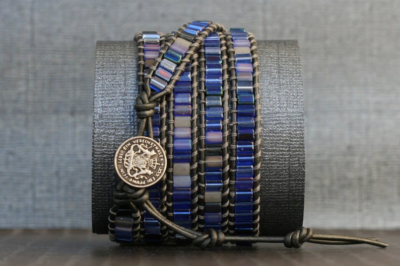 Puede incluir: Un brazalete de cuero de m&uacute;ltiples hebras con cuentas de vidrio azul y gris y un dije de moneda de plata. El brazalete est&aacute; envuelto alrededor de un cilindro negro.