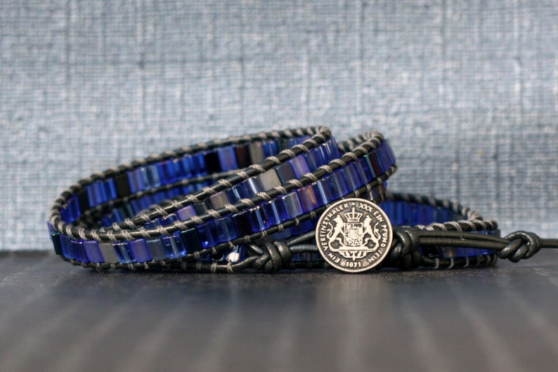 Puede incluir: Un brazalete de envoltura de cuentas azules y negras con un dije de moneda de plata que presenta un escudo de armas.
