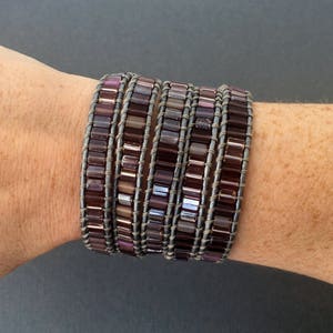 May include: A multi-layered bracelet made of small, square-shaped beads in shades of purple and gray. The bracelet is wrapped around a person's wrist.