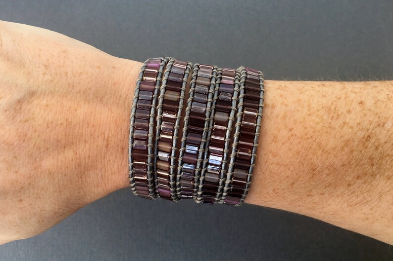 May include: A multi-layered bracelet made of small, square-shaped beads in shades of purple and gray. The bracelet is wrapped around a person's wrist.