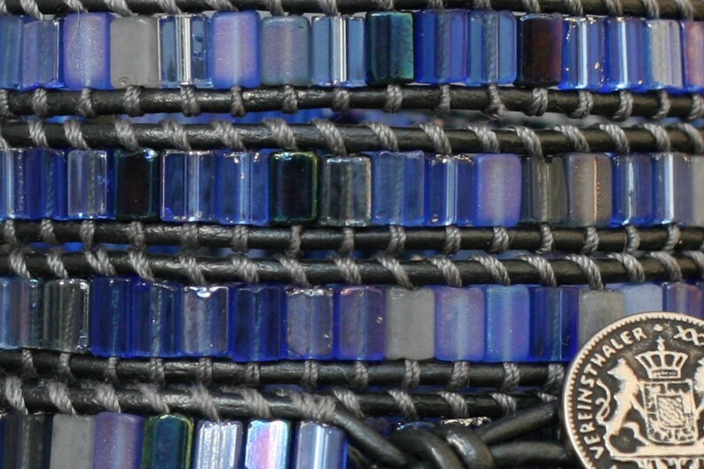 Puede incluir: Un primer plano de una pulsera de cuentas azules y grises con un dije de moneda de plata. La moneda tiene las palabras "Vereinsthaler" y un escudo de armas.