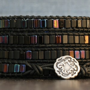 May include: Black leather wrap bracelet with iridescent black square beads and a silver charm with a swirling design.