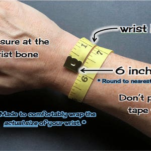 May include: A yellow measuring tape wrapped around a wrist to show how to measure for a bracelet. The text reads "Measure at the wrist bone", "wrist bone", "6 inches", "*Round to nearest half inch*" and "Don't pull the tape tight".