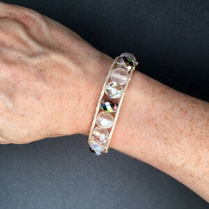 May include: A white beaded bracelet with iridescent crystal beads. The bracelet is made with a white cord and is worn on a person's wrist.