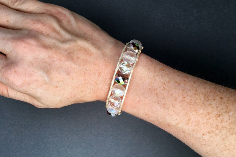 May include: A white beaded bracelet with iridescent crystal beads. The bracelet is made with a white cord and is worn on a person's wrist.