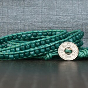 May include: A teal wrap bracelet with wooden beads and a silver button clasp. The bracelet is made of leather and wraps multiple times around the wrist. The clasp has an engraved design.