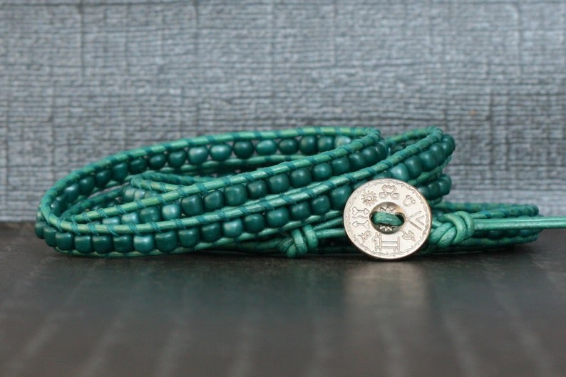 May include: A teal wrap bracelet with wooden beads and a silver button clasp. The bracelet is made of leather and wraps multiple times around the wrist. The clasp has an engraved design.