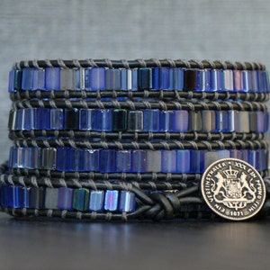 Puede incluir: Un brazalete de cuero negro con m&uacute;ltiples filas de cuentas de vidrio azul iridiscente. El brazalete est&aacute; atado con un nudo y tiene un dije de moneda de plata con un escudo de armas y el texto "EIN EIN PFUND 1871".