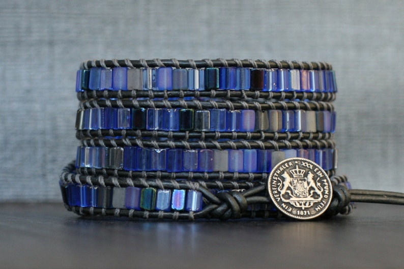 Puede incluir: Un brazalete de cuero negro con m&uacute;ltiples filas de cuentas de vidrio azul iridiscente. El brazalete est&aacute; atado con un nudo y tiene un dije de moneda de plata con un escudo de armas y el texto "EIN EIN PFUND 1871".