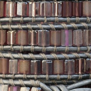 May include: A close-up of a bracelet made of leather and small, rectangular, iridescent glass beads. The beads are arranged in rows and are a mix of purple, pink, and brown.