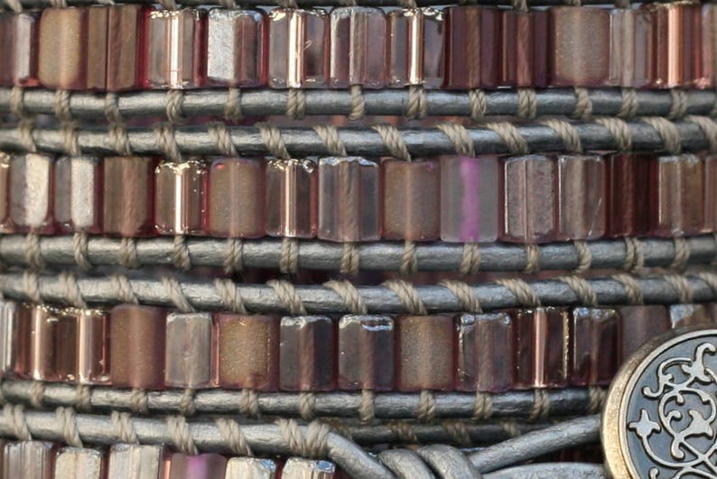 May include: A close-up of a bracelet made of leather and small, rectangular, iridescent glass beads. The beads are arranged in rows and are a mix of purple, pink, and brown.