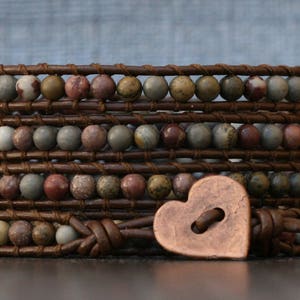 May include: A multi-layered leather bracelet with brown leather straps and colorful stone beads. A copper heart-shaped charm with a button detail hangs from the bracelet.