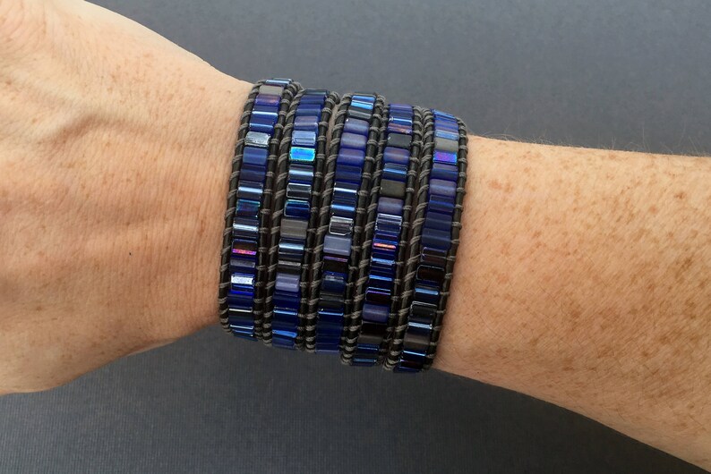 Puede incluir: Un brazalete de m&uacute;ltiples capas con cuentas de vidrio azul y negro iridiscentes y una base de cuero marr&oacute;n.